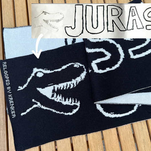 Deze zwart-witte kindersjaal heeft een ontwerp met een dinosaurus. De witte dinosaurus is gebreid op een zwarte achtergrond.